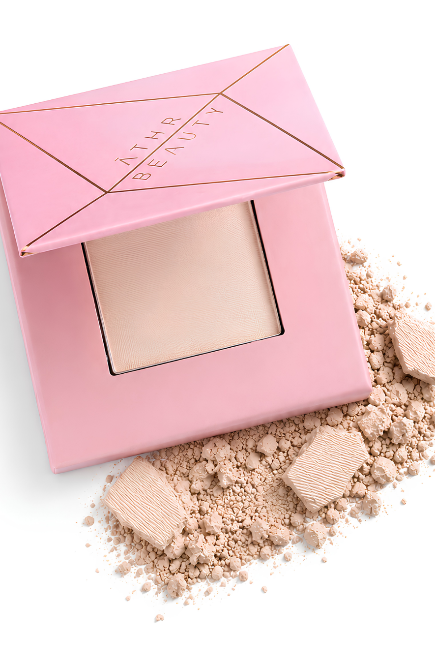 Athr Beauty's pink makeup compact of the 'Desert Mirage Blurring Powder' with open lid and scattered powder in the shade 'Rose Mesa' (a soft, creamy beige with a subtle pink tint) on a white background